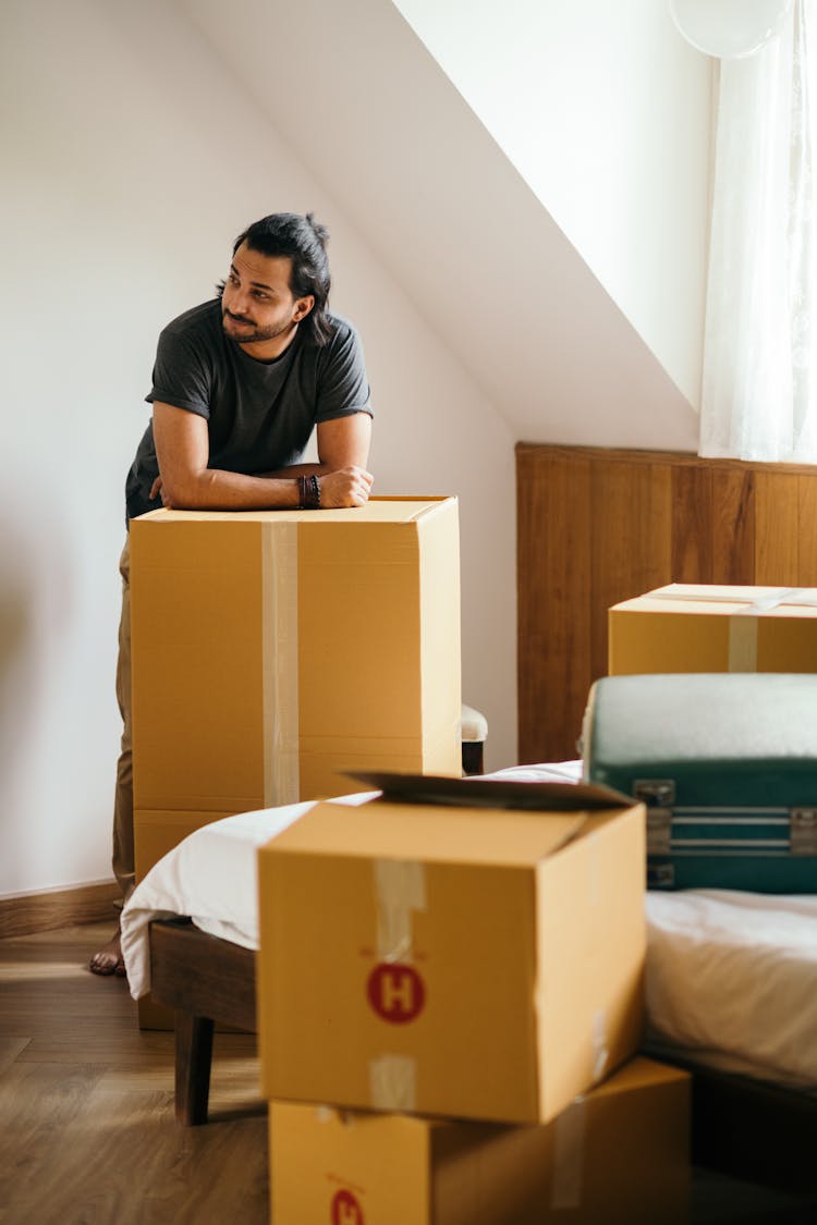 Thoughtful Ethnic Man Standing In New House Leaning On Unpacked Carton Box