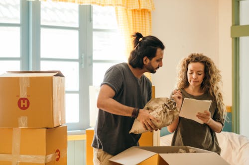 Free Cheerful young man and woman smiling while unpacking carton boxes with belongings in new apartment during relocation and looking at paper Stock Photo