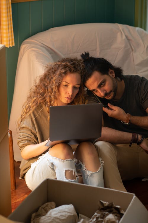Concentrated couple sitting on floor near armchair and browsing laptop while spending time together and searching information about relocation at night