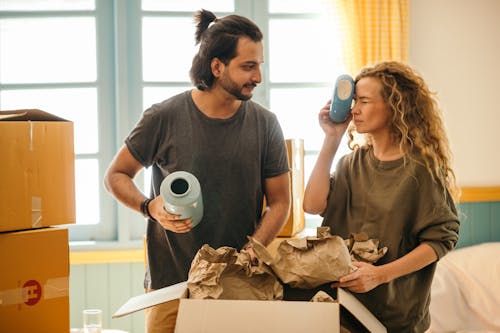 Satisfied adult bearded ethnic man and playful woman with curly hair wearing casual t shirts unboxing ceramics for home decoration from carton boxes while moving in new apartment