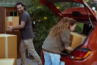 Young woman with curly hair getting carton box out from trunk of automobile while cheerful ethnic man carrying box into new home in suburb or countryside area