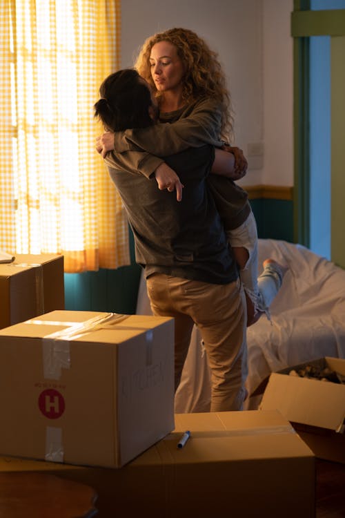 Young man lifting attractive woman with curly hair while standing in living room among carton boxes and chair covered with white fabric while starting living together in new apartment