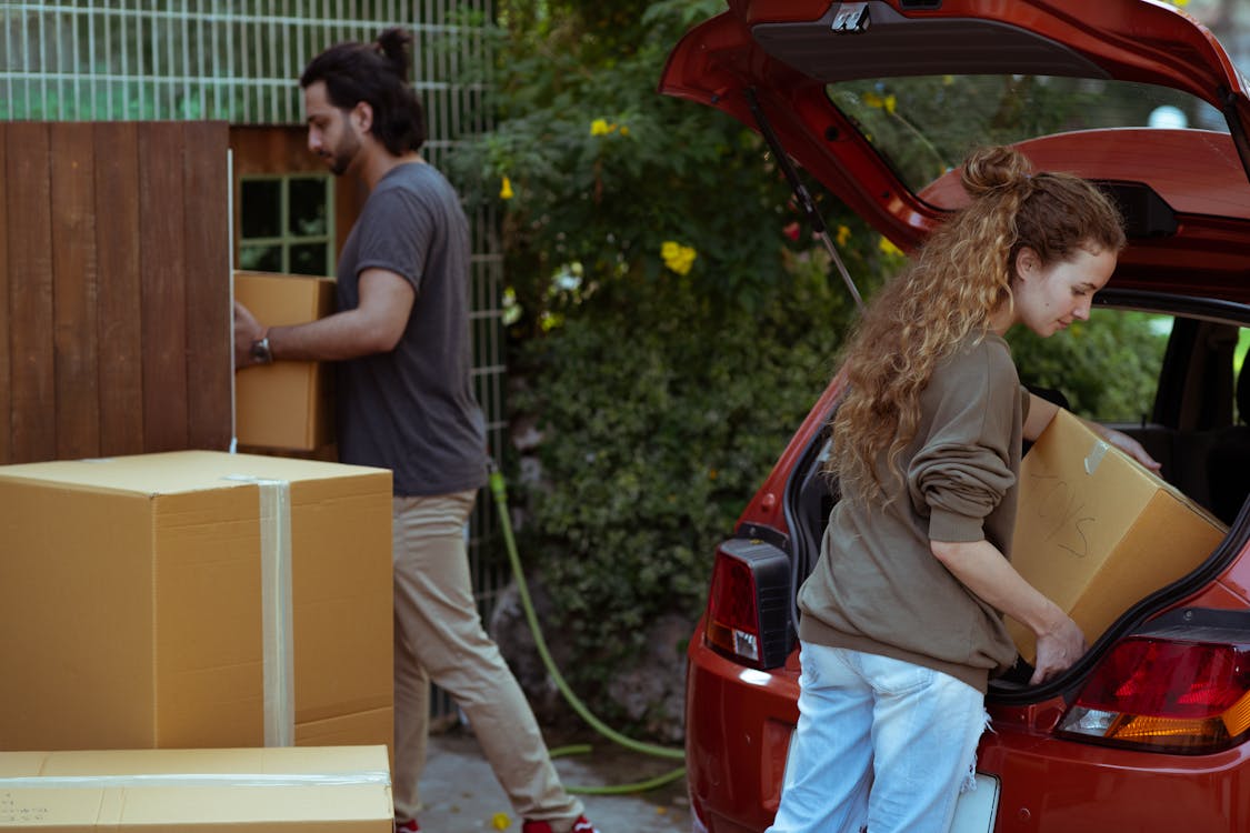 Free Focused young woman with curly hair taking cardboard package out from red automobile trunk on street near stack of moving boxes while bearded ethnic man carrying box into new house Stock Photo car last longer