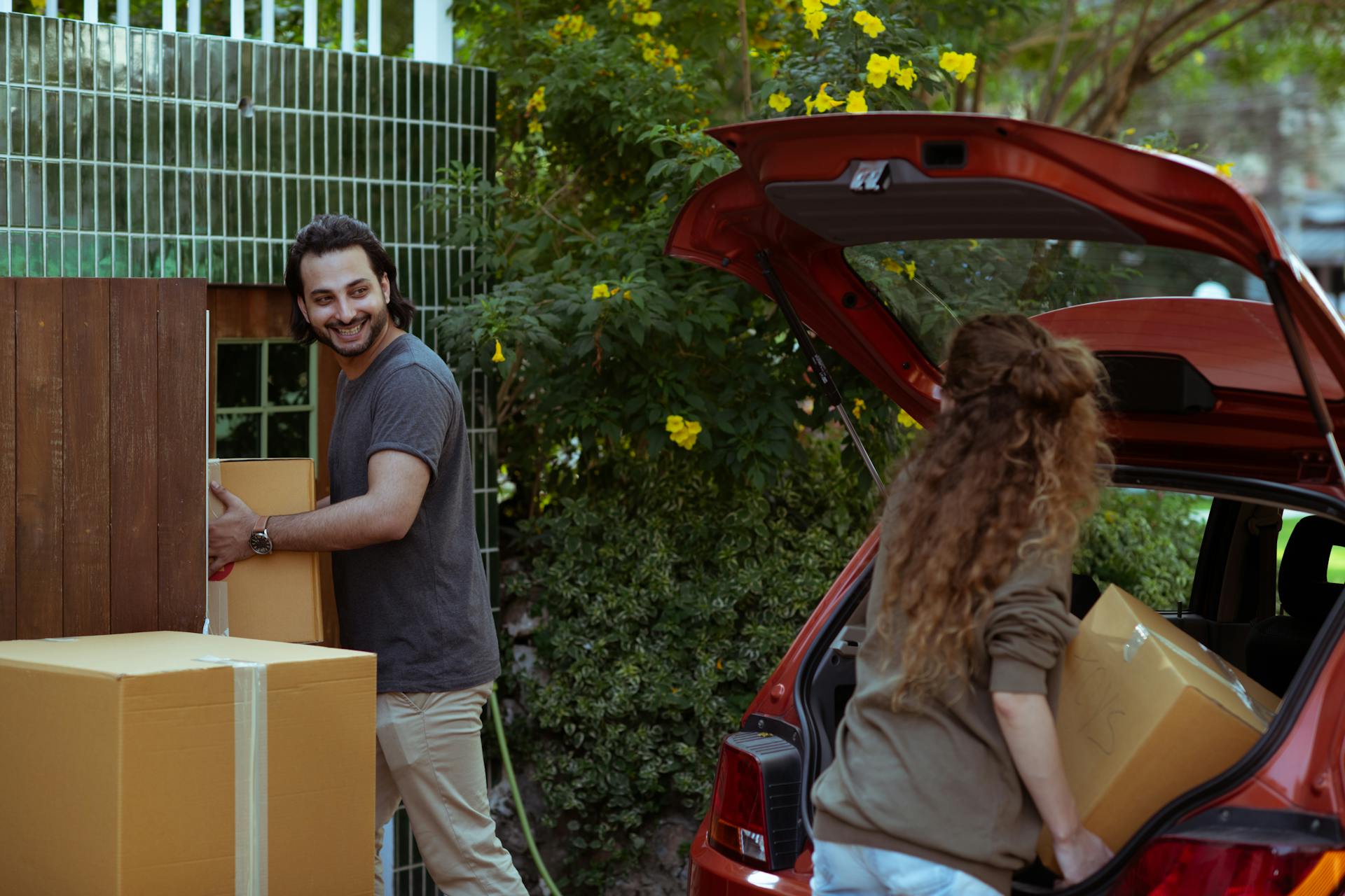 Happy couple in casual clothes moving personal items in carton boxes from car trunk to new house