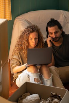 From above of concentrated couple in casual clothes sitting on floor among carton boxes and covered with sheet arm chair while using netbook and calling by mobile phone