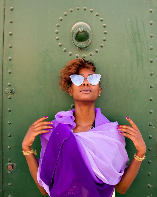 Young African American female in trendy purple organza scarf and sunglasses with modern hairstyle touching shoulders while looking up and standing near green door