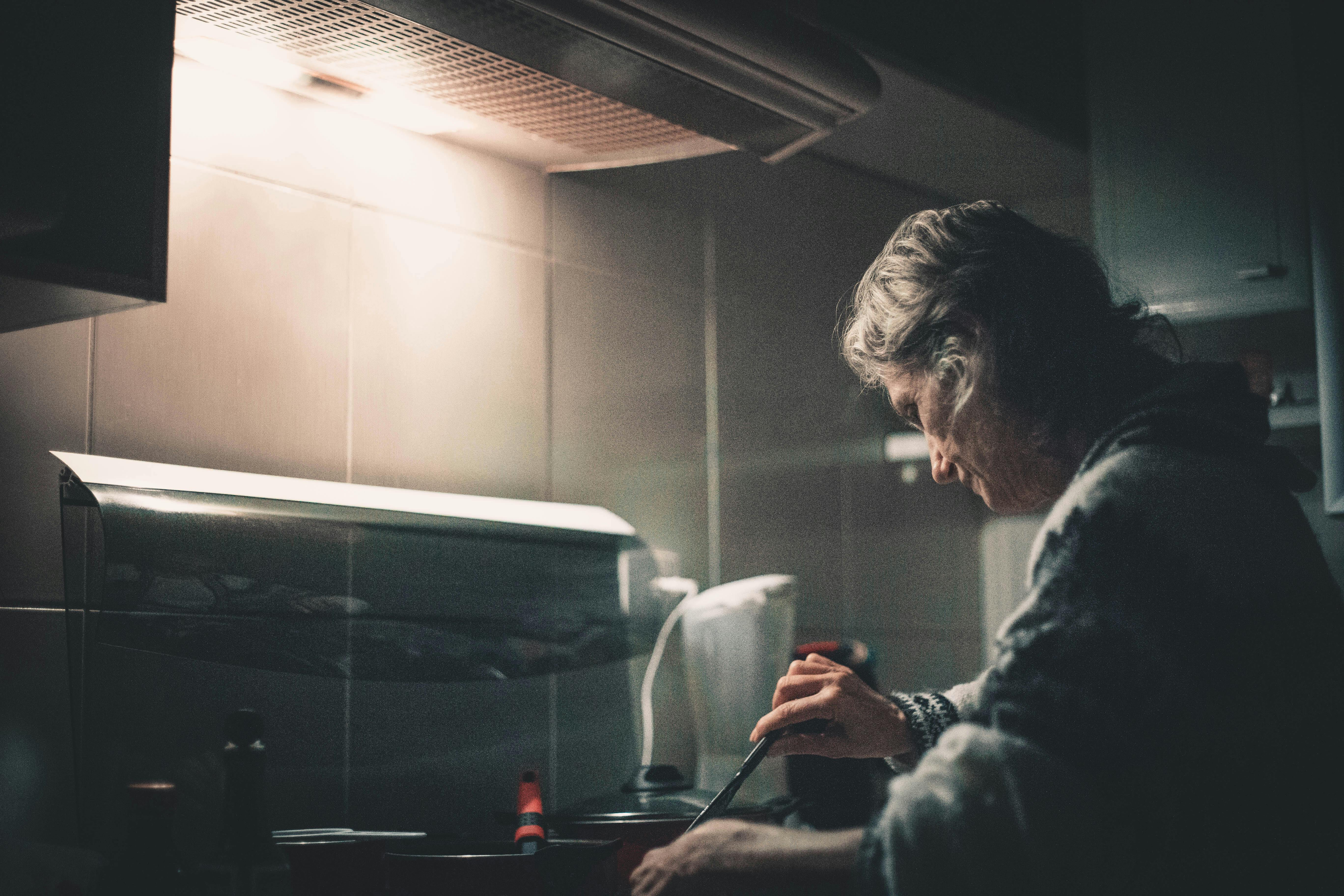 Senior woman preparing food in a dimly lit kitchen, creating a warm and homey atmosphere.