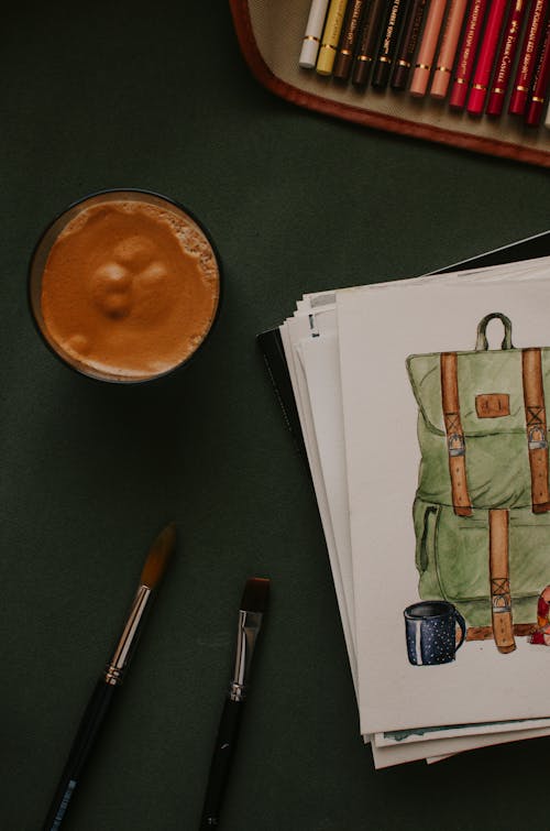 Top view of sketchbook with colorful picture of backpack placed near brushes and various colored pencils
