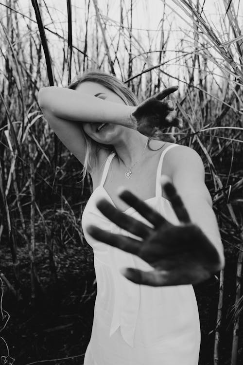 Black and white of cheerful young female in casual white dress smiling and covering face with dirty palms in summer field