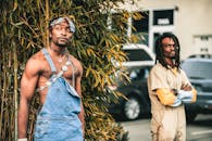 Angry African American male in work uniform standing on street with arms folded and looking enviously at brutal fit informal black man with naked torso