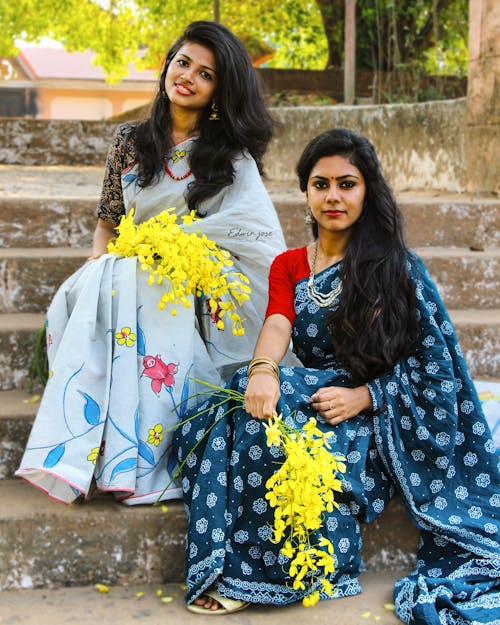 Full length young cheerful Indian ladies with long voluminous hair wearing traditional authentic sari dresses sitting on street steps with yellow flowers