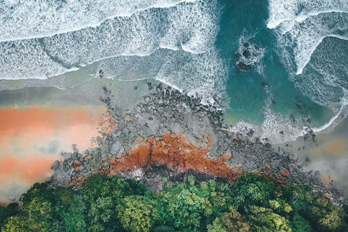 Drone view of turquoise foamy sea waves rolling calmly near stony shore with lush greenery in exotic island