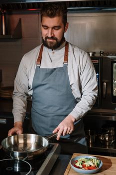 Chef in apron cooking with skillet, preparing vegetables in a modern kitchen setting.