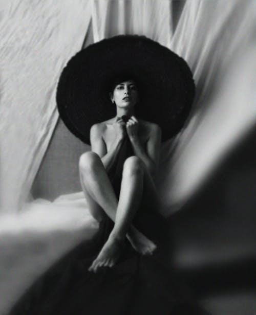 Free Black and white of gorgeous barefooted young female in elegant hat sitting on floor with crossed legs and covering body with cloth Stock Photo