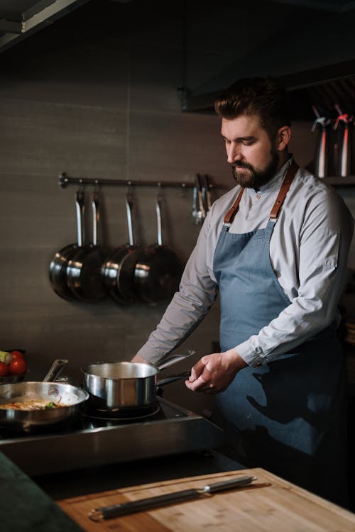 Man in White Dress Shirt and Blue Apron Holding Stainless Steel Cooking Pot