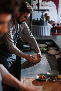 Chef kneading dough on a metal table in a modern open kitchen setting.