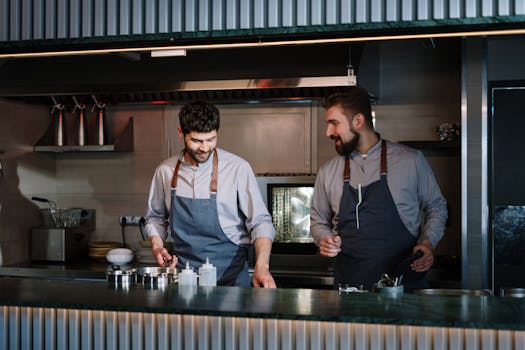 Two chefs collaborate in a sleek, industrial kitchen space focused on creative culinary arts.