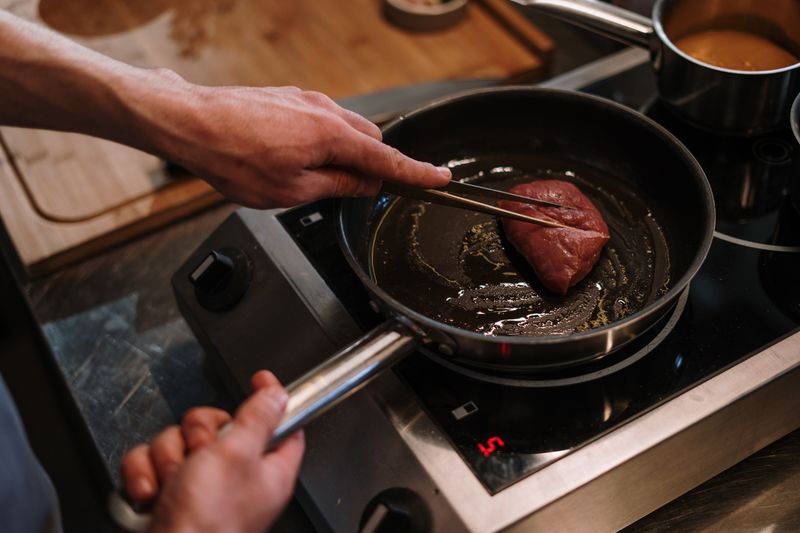 A chef sears a steak in a skillet on an induction cooktop in a professional kitchen.