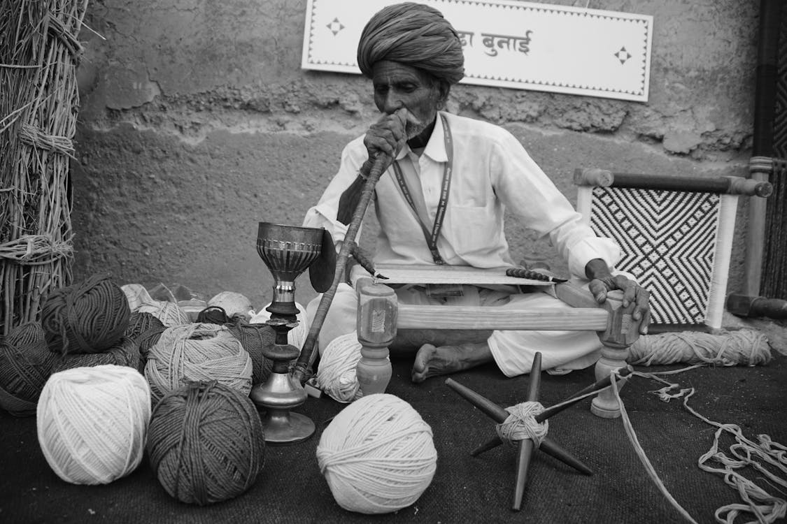 Free Black and white of senior barefooted Indian male in skirt and turban smoking hookah while weaving traditional carpet on authentic wooden machine on street Stock Photo