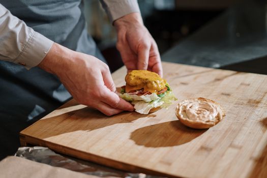 Chef crafting a delicious cheeseburger with lettuce, tomato, and cheese on a wooden cutting board indoors.