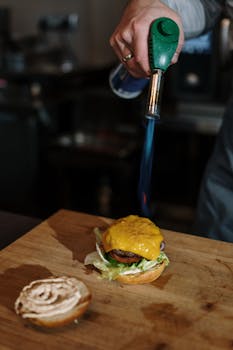Chef uses a torch to perfectly melt cheese on a gourmet burger in a professional kitchen.