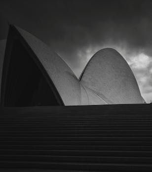 Low angle of black and white stairs and details of sail shaped roofs of contemporary Sydney Opera House against overcast sky