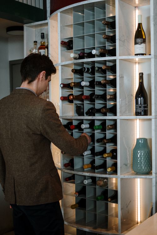 Free Man in a brown blazer choosing wine from a modern wine rack in a restaurant setting. Stock Photo