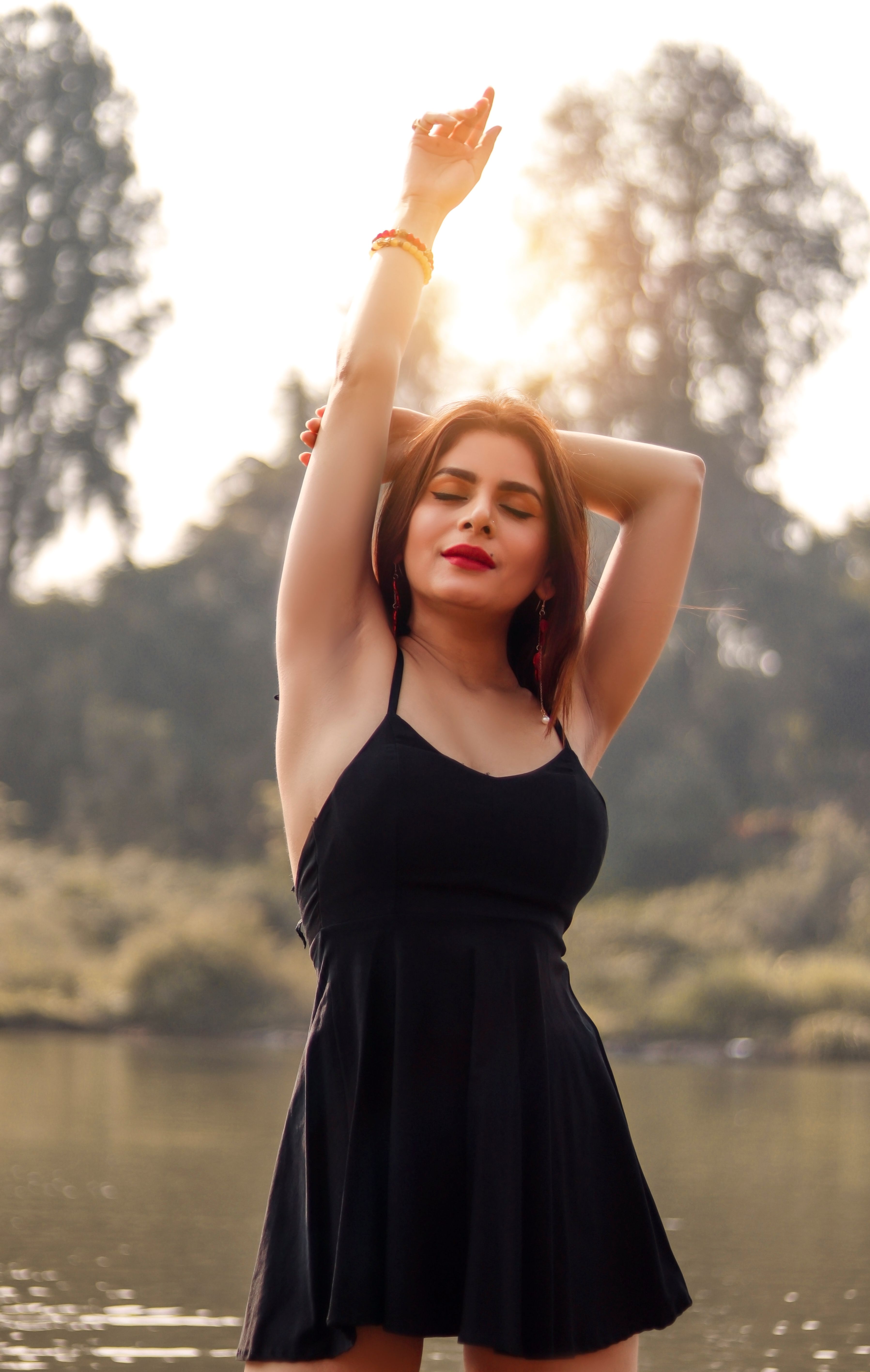 Elegant woman in black dress with arms raised, enjoying a peaceful outdoor moment at sunset.