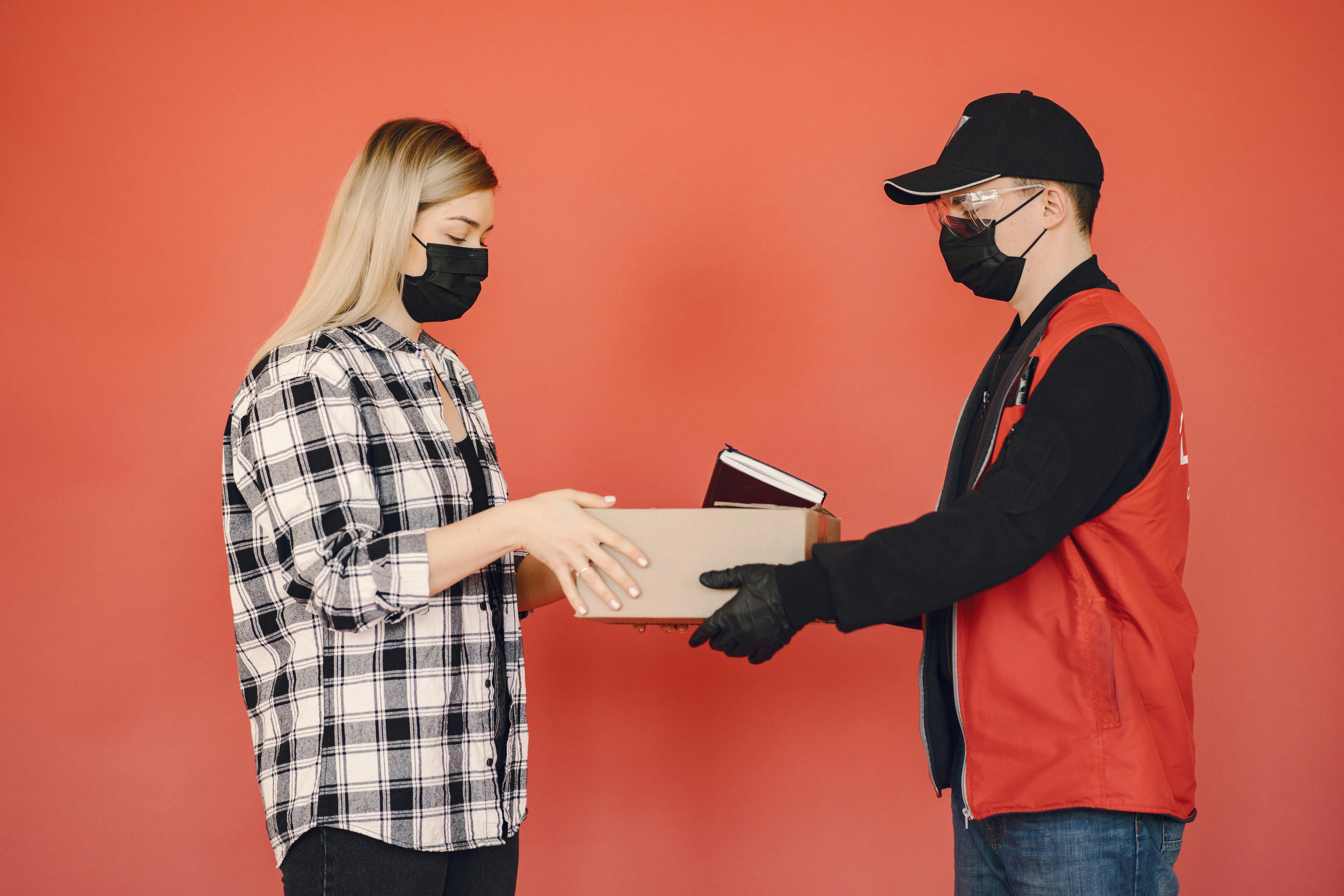 Side view of adult delivery man in protective mask and goggles with gloves giving carton parcel to woman in checkered shirt and medical mask isolated on red background illustrating concept of safety delivery during quarantine by Gustavo Fring