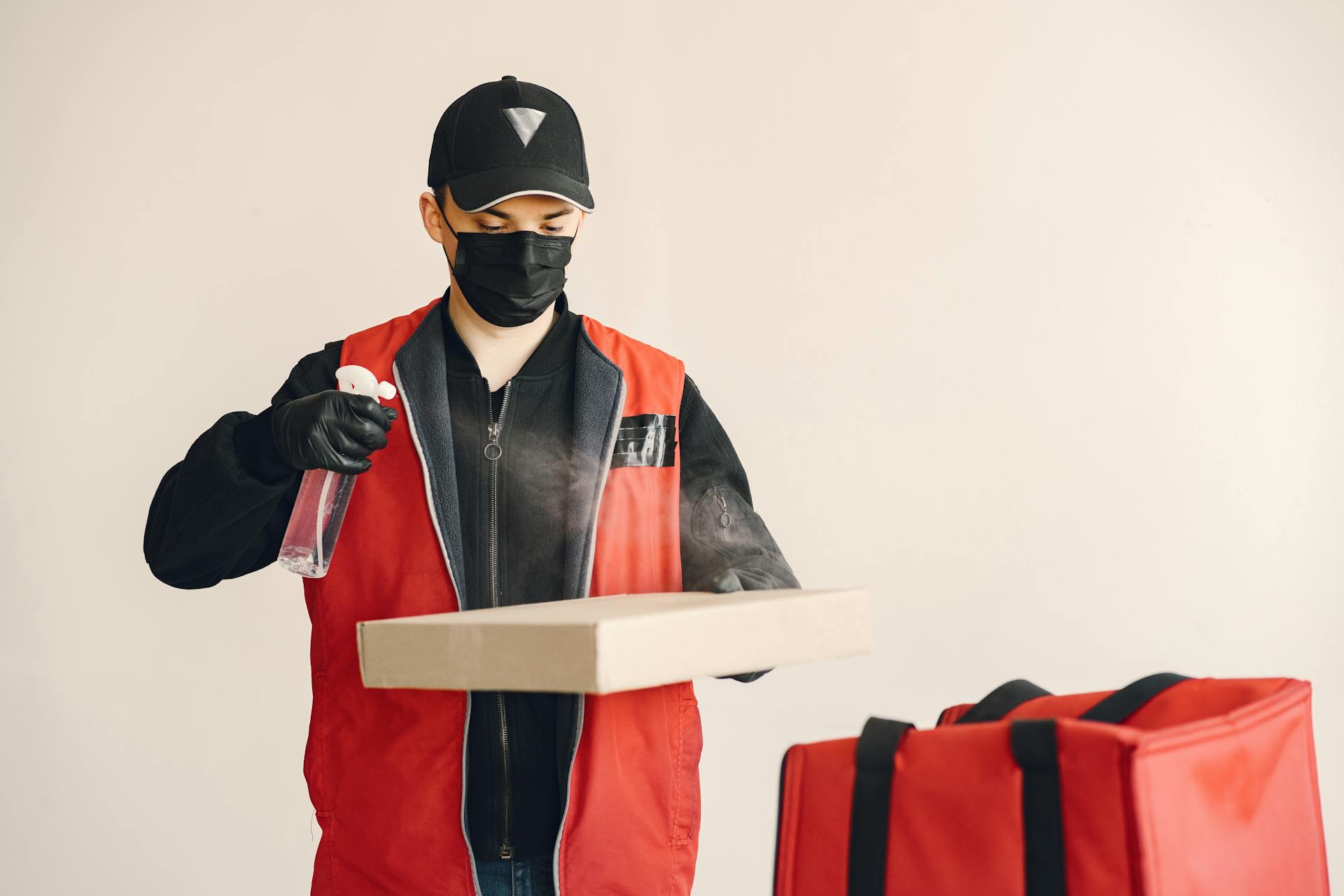 Male courier in cap and uniform wearing medical facial mask and gloves spraying antiseptic on carton pizza box against white background near thermo bag illustrating safety food delivery concept