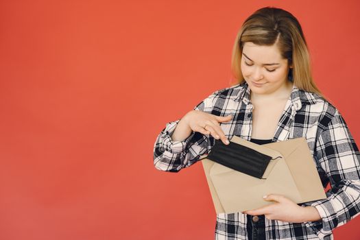 Happy female in plaid shirt taking surgical protective mask out from craft paper envelope isolated on red background illustrating safety mail deliveries during coronavirus pandemic