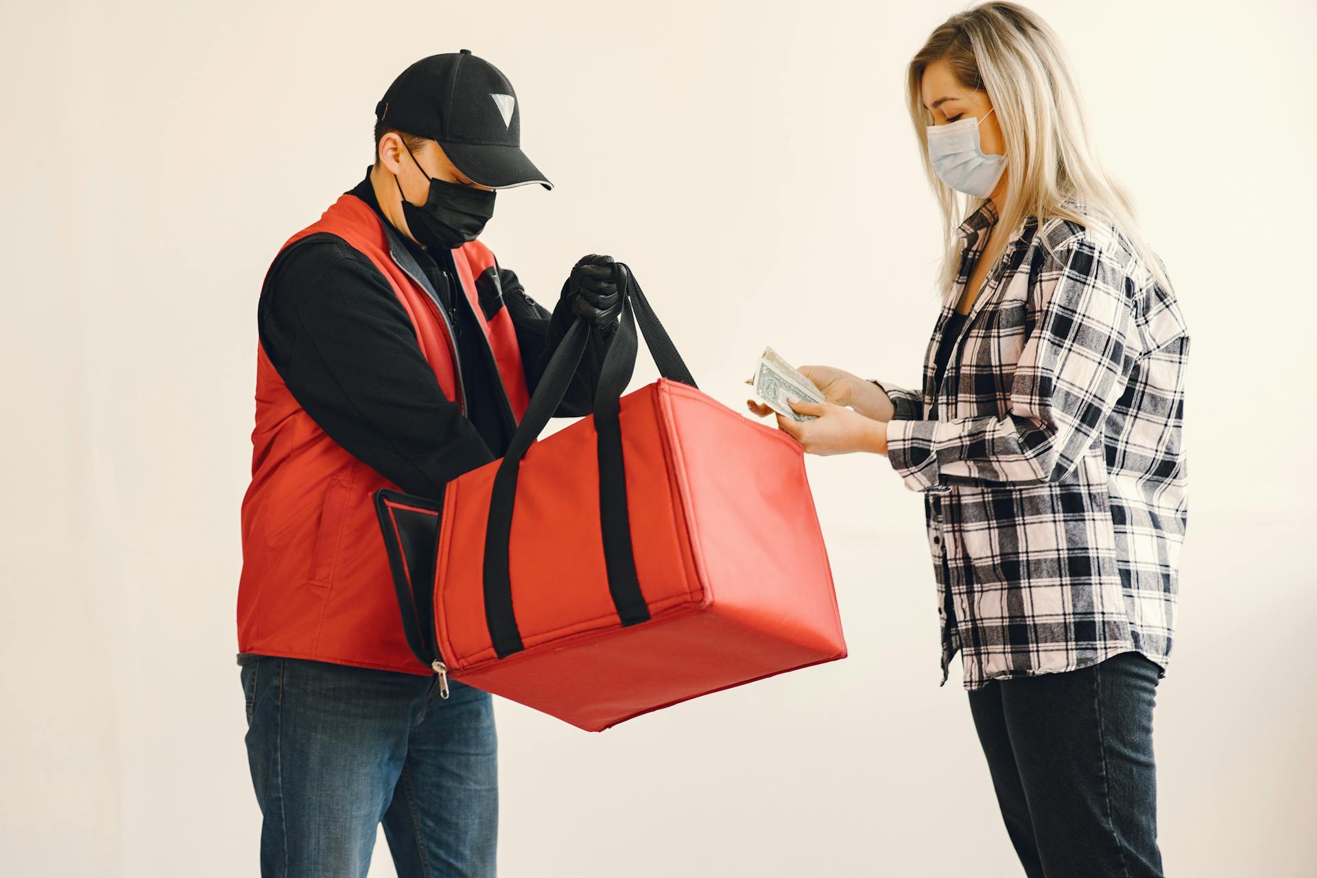 Young blond woman in checkered shirt and medical mask paying cash to young delivery man in protective mask and gloves during safety food delivery in studio