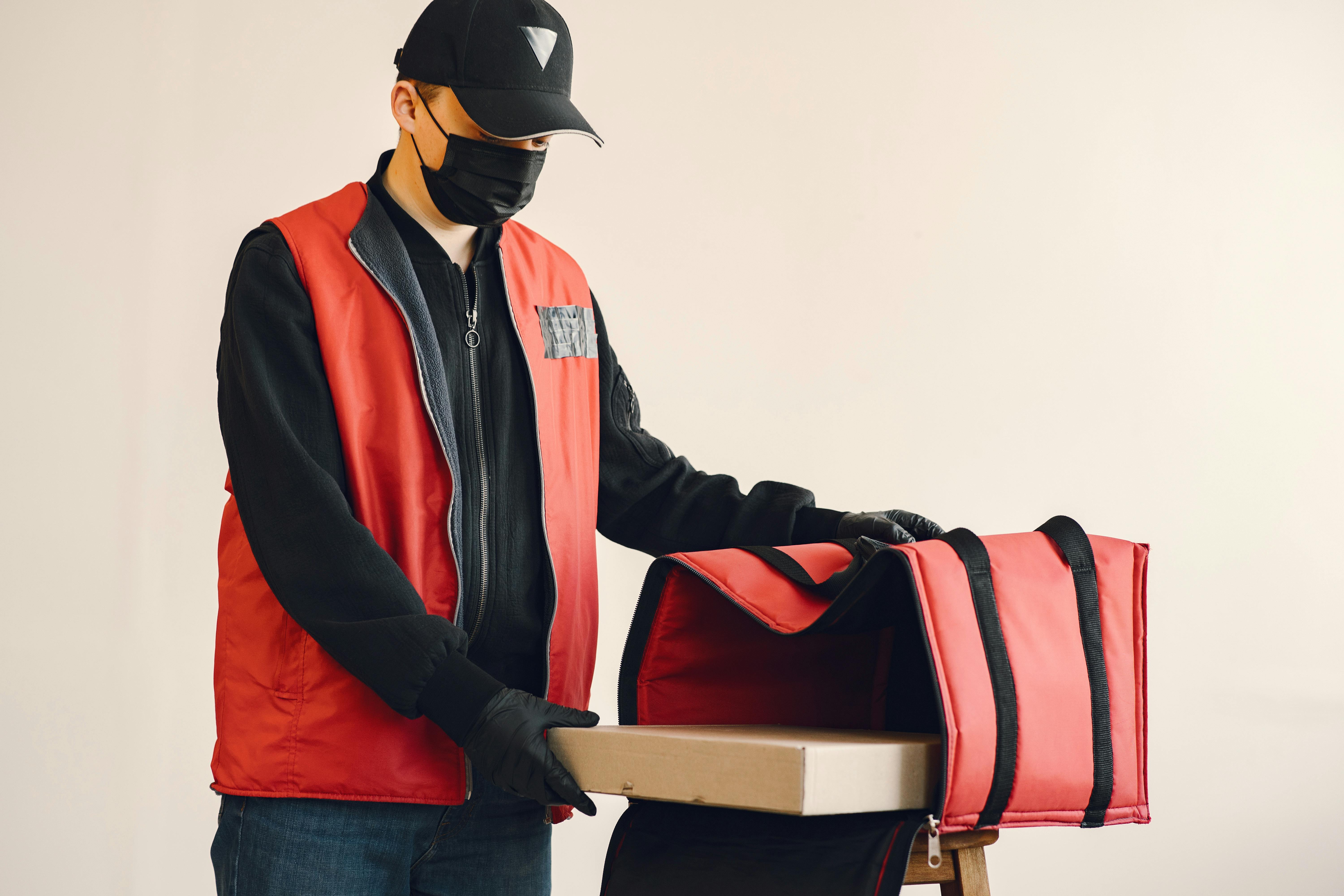 male courier in uniform and medical mask with thermo bag and pizza in studio