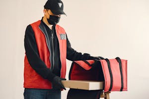 Young delivery man wearing surgical facial mask and gloves for preventing disease during coronavirus pandemic putting pizza box in thermo bag for safety food delivery