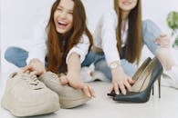 Crop exited young female in jeans and white shirts choosing between elegant glamour black high heels and comfortable trendy suede sneakers while sitting on floor together