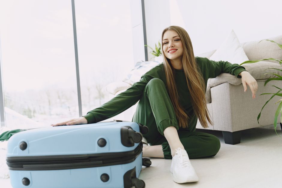 Young woman in green tracksuit smiling with suitcase in modern living room, ready for travel.