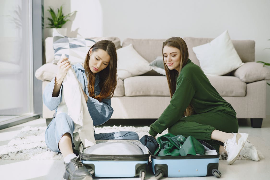 Free Optimistic young female travelers in trendy outfits sitting on floor against comfortable sofa and unpacking suitcase in modern hotel room Stock Photo