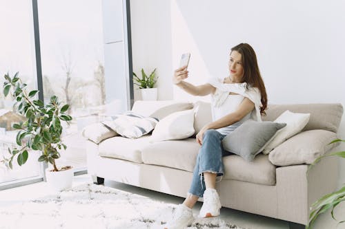 Positive young female doing selfie on smartphone while sitting on sofa
