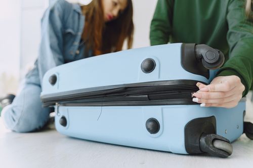 Free From below of crop concentrated young woman in denim outfit closing suitcase sitting on floor with anonymous friend while preparing for trip Stock Photo