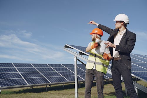Young businessman in formal outfit and foreman in yellow vest looking away while discussing working process against solar panels