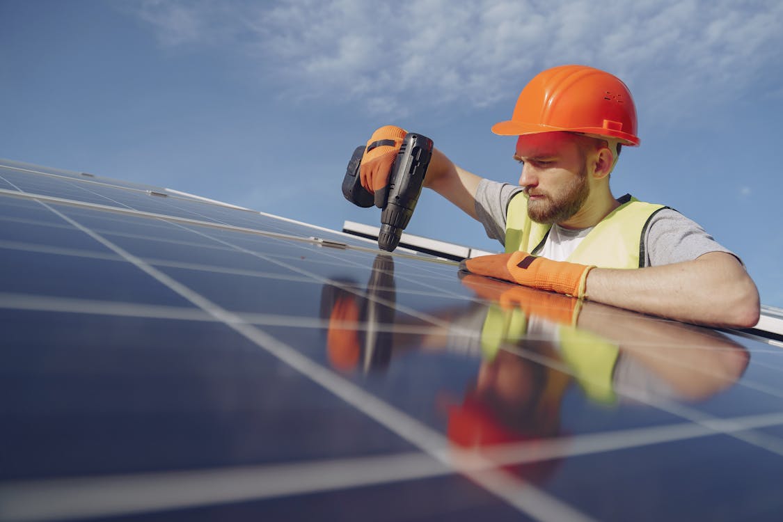Free Low angle of serious bearded male electrician in hardhat and protective gloves installing solar photovoltaic panel system using drill Stock Photo
