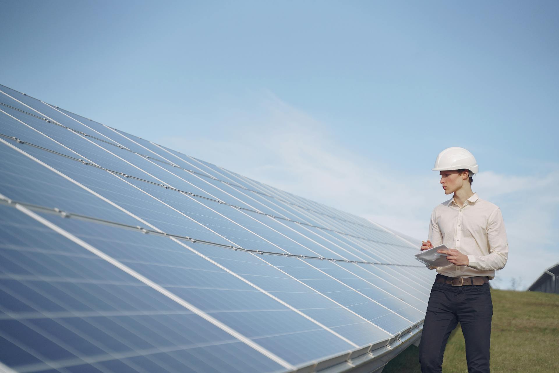 Young serious male engineer in formal outfit and helmet reporting while inspecting solar panel field