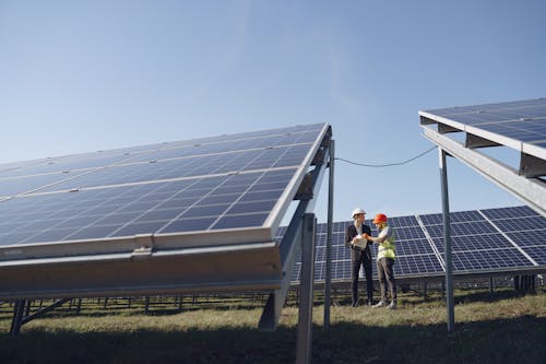 Supervisor in hardhat and formal wear discussing project with workman in uniform while standing with papers near modern solar panels under blue sky