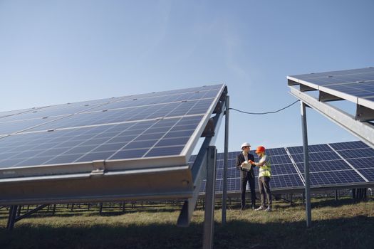 Photo by Gustavo Fring Supervisor in hardhat and formal wear discussing project with workman in uniform while standing with papers near modern solar panels under blue sky