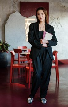 一位时尚的年轻女子在咖啡馆里，手拿笔记本，自信地站在室内