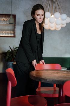 A woman in a black suit arranges seating in a modern cafe with red chairs and chic lighting.