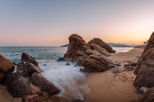 Free Magnificent scenery of huge rough stones on sandy beach of waving ocean against cloudless sunset sky Stock Photo