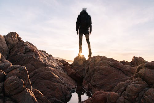 Free Full body unrecognizable tourist in black jacket with backpack standing on rocky shore and admiring sunrise Stock Photo