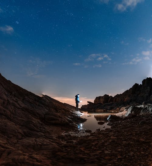 Unrecognizable tourist in warm clothes standing on rocky coast near water puddle against evening sky