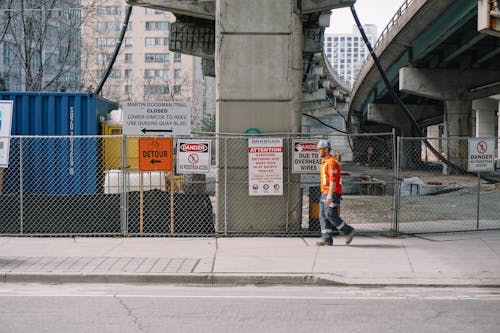 Full body side view of adult male worker in orange uniform and grey helmet walking under big bridge along chain link fence on street in daylight